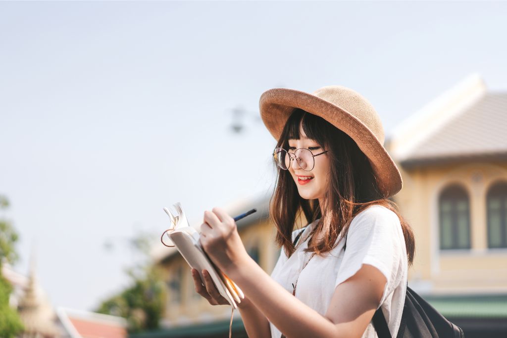 A young woman in a straw hat and glasses writing in a notebook while exploring a scenic town, symbolizing adventures in backpacking Southeast Asia and documenting Southeast Asia experiences.