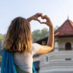 Woman making a heart shape with her hands in front of a Sri Lankan temple, wearing a white top and blue shawl. Highlights the best time to visit Sri Lanka for a cultural experience.