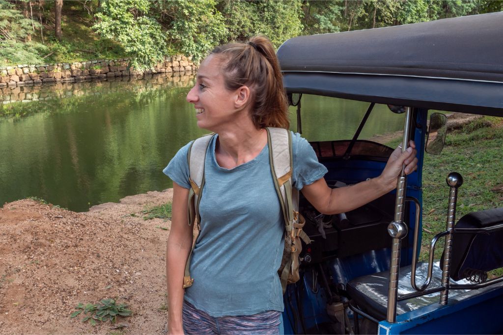 A woman dressed in a casual outfit with a backpack poses near a tuk-tuk by a tranquil river. Ideal for Sri Lanka travel outfit ideas and tips on packing for Sri Lanka.