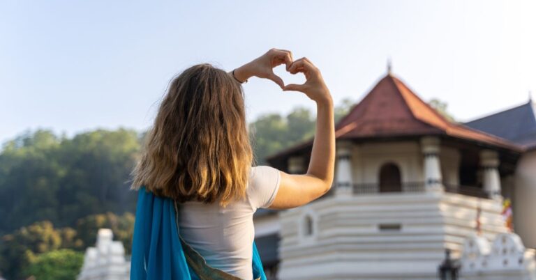 Woman making a heart shape with her hands in front of a Sri Lankan temple, wearing a white top and blue shawl. Highlights the best time to visit Sri Lanka for a cultural experience.