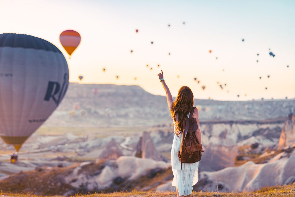 A woman with a backpack points at colorful hot air balloons over the stunning rock formations of Cappadocia, Turkey, a dreamy addition to a 2025 travel bucket list and Asia bucket list travel goals.