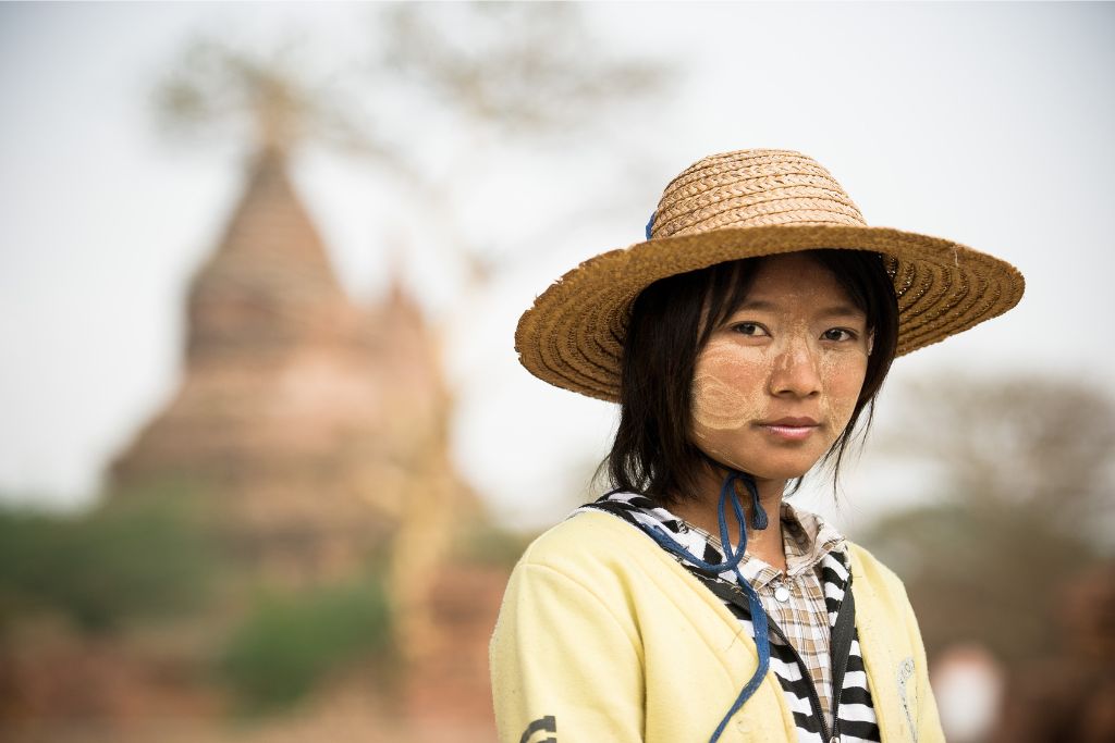 A young woman with traditional Thanaka face paint and a straw hat poses near ancient temples in Myanmar, offering a glimpse into unique Southeast Asia experiences and hidden gems in Asia.