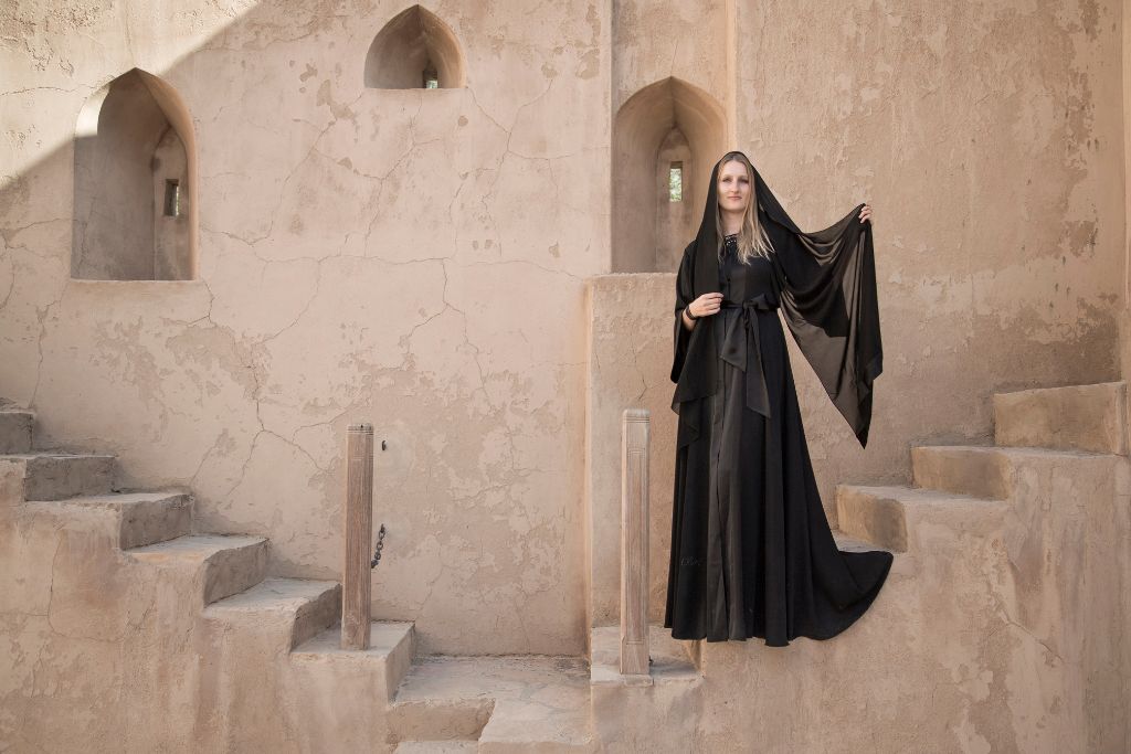 A woman wearing a black abaya poses gracefully against an ancient mud-brick fortress with arched windows in Oman. Known for its rich heritage and desert warmth, Oman is an ideal warm winter destination in Asia, offering history, architecture, and sunshine for travelers escaping the cold.