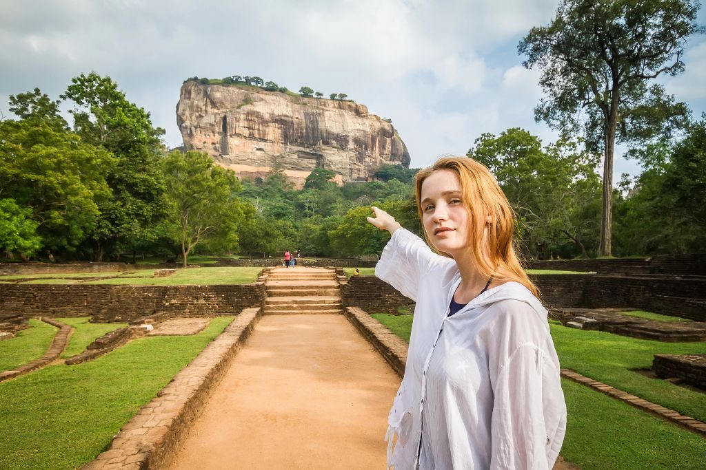 A traveler in a white hooded shirt points to the towering Sigiriya Rock Fortress in Sri Lanka, set against lush greenery and ancient ruins. Sri Lanka is one of the best places to visit in Asia in December for history, hikes, and tropical weather that feels worlds away from winter.