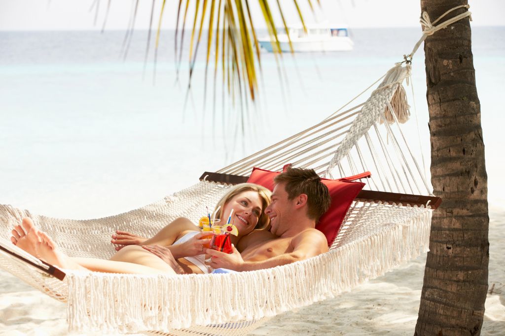 A couple lounges in a woven hammock strung between palm trees on a white sand beach in the Maldives, holding colorful tropical drinks. The Maldives is a luxury winter getaway in Asia, perfect for honeymooners and sun-seekers wanting crystal-clear waters and warm ocean breezes.