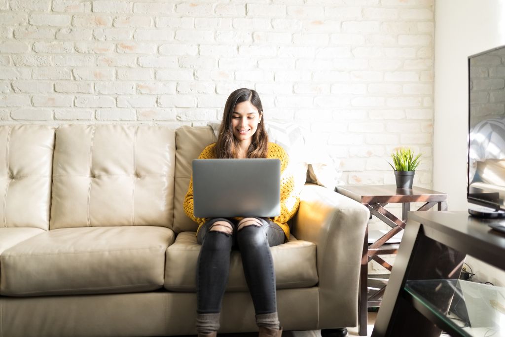 A woman sitting on a beige couch using a laptop, planning her travel itinerary and learning tips on how to haggle while traveling abroad