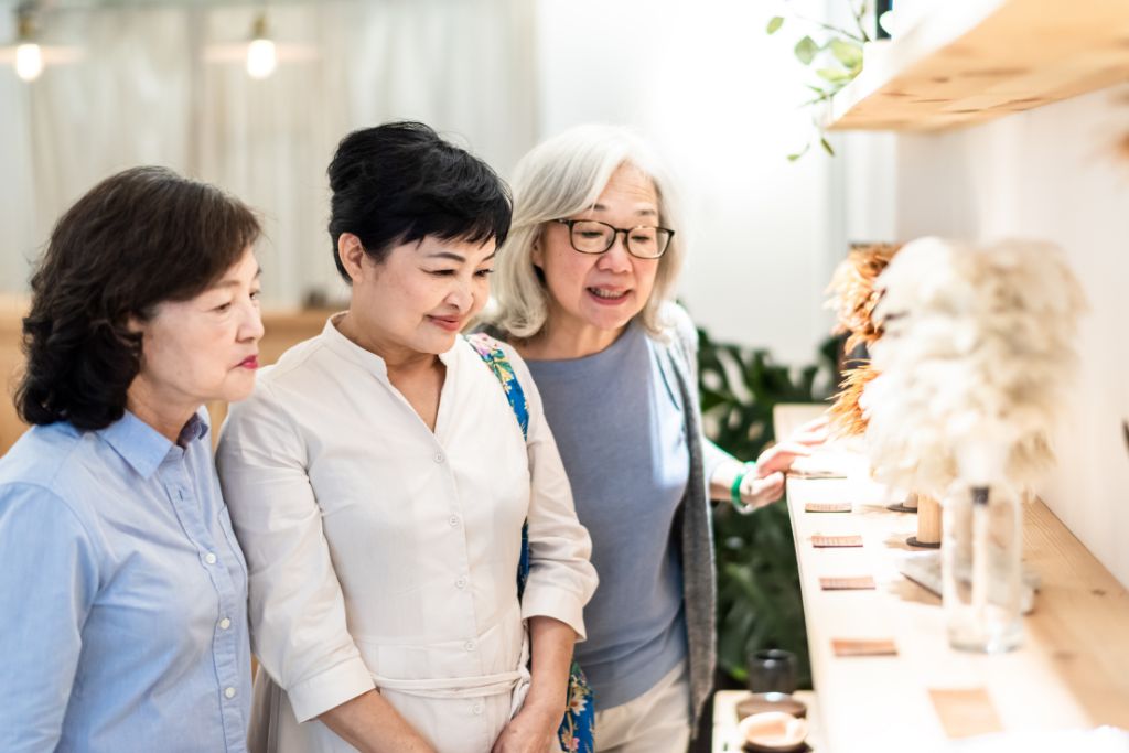 Three older women smiling as they examine handmade crafts at a market, highlighting the cultural experience of bargaining in countries like Mexico, Thailand, or Turkey.