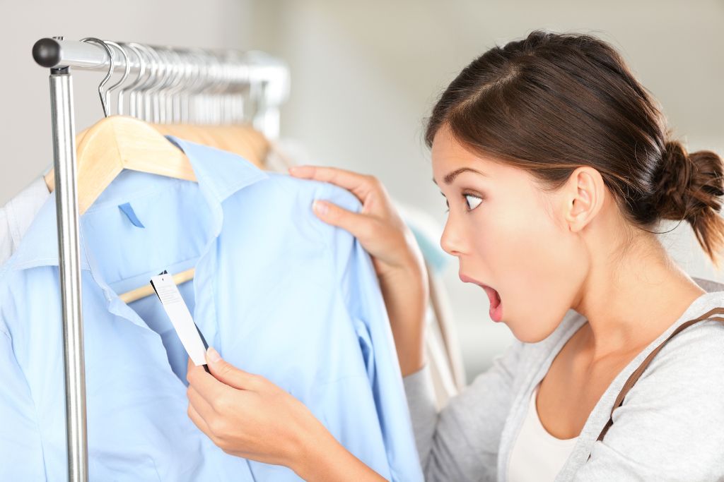 A surprised woman holding up a shirt in a store, reacting to the price tag, an ideal moment to use haggling tips for negotiating prices.