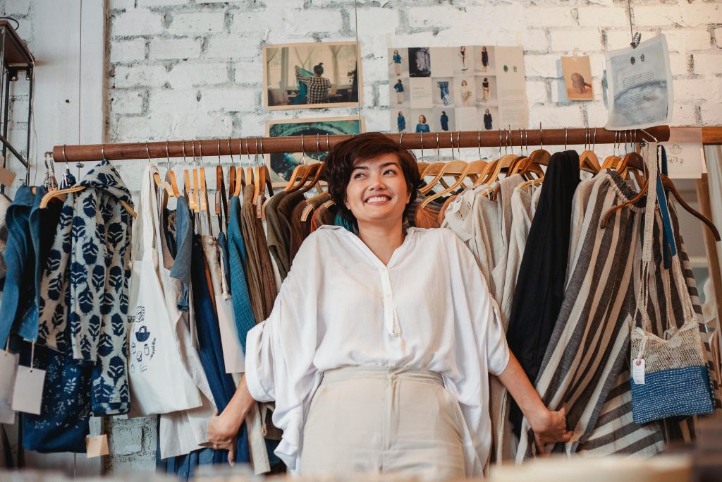 A woman in a white blouse stands among racks of clothing, smiling as she playfully grabs garments on both sides. Knowing how to bargain in a boutique or market can make shopping more fun and affordable. Use smart haggling tips to get the best deal.