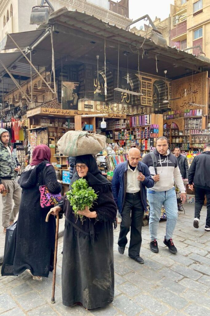 A local woman carrying fresh herbs on her head in Khan el Khalili, Cairo, against the backdrop of a traditional spice and handcrafts shop, representing everyday life in the Cairo marketplace.