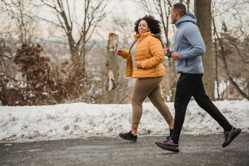 : A woman in a yellow jacket and a man in a hoodie jog on a snowy path, smiling. Staying fit while traveling is great, but don’t forget to haggle in the market for the best deals. Stop overpaying and negotiate smartly.