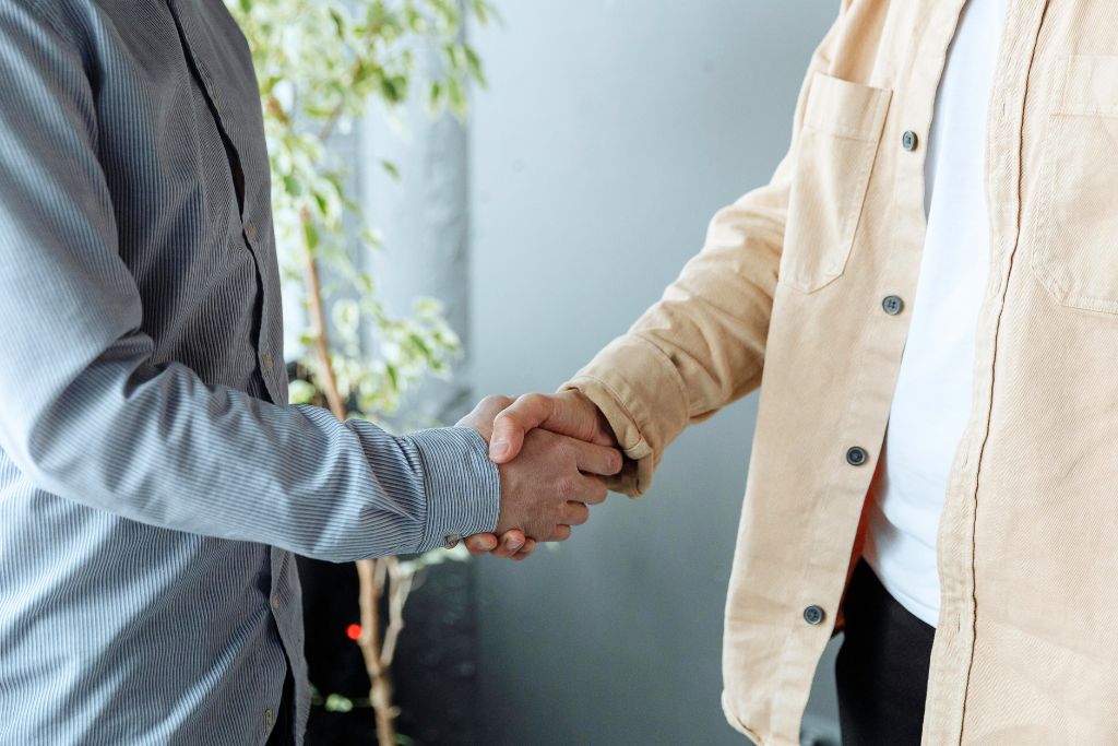 A close-up of two people shaking hands, one in a beige jacket and the other in a striped shirt. A successful bargaining agreement is sealed with a handshake. Haggling etiquette is key to a fair deal.