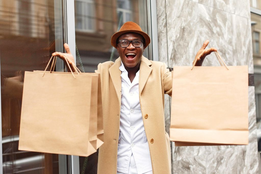 A man in a camel coat and hat excitedly holds up large shopping bags outside a store. This is the result when you ask to lower the price successfully. The best way to haggle abroad is to start with a friendly approach.