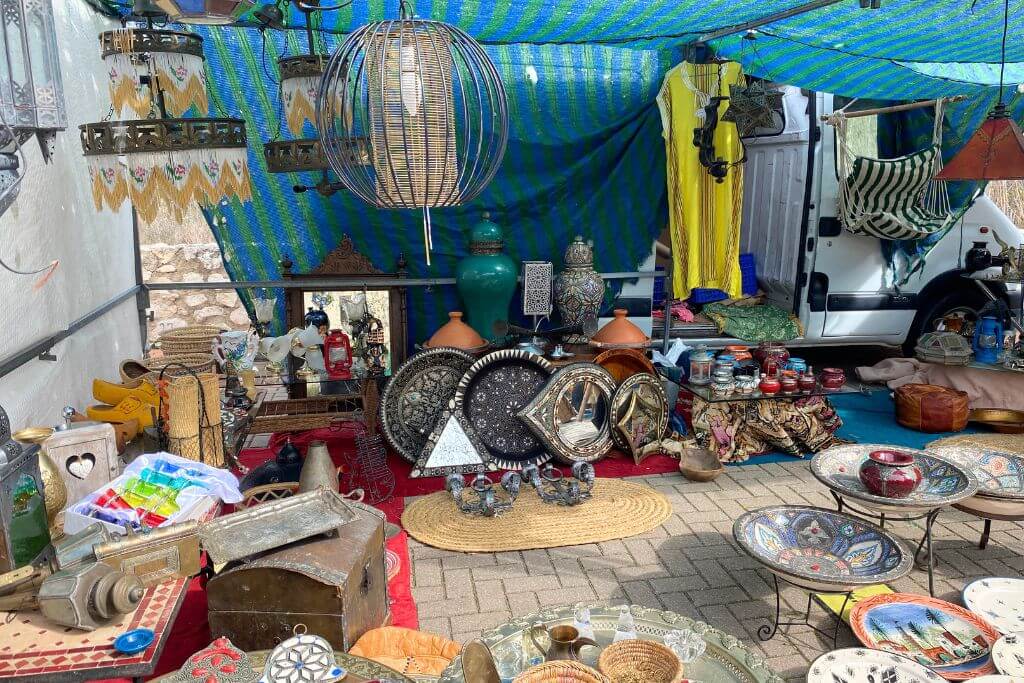 A colorful market stall under a striped canopy displays Moroccan lamps, ceramic plates, and decorative items. This is a great place to haggle in the market and practice negotiation etiquette for a fair deal.