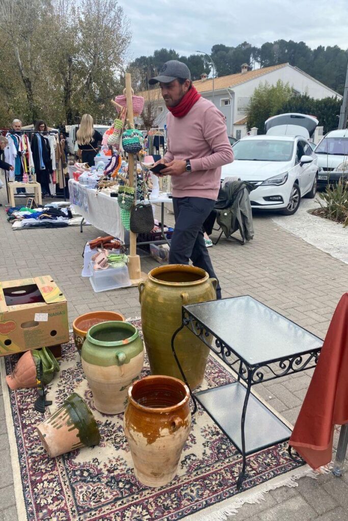 A man in a red scarf and cap walks past large clay pots and woven baskets at an outdoor flea market. Mastering how to bargain while on holiday can help you negotiate fair prices on handcrafted goods. Vendors expect bartering, so don’t hesitate to start low.