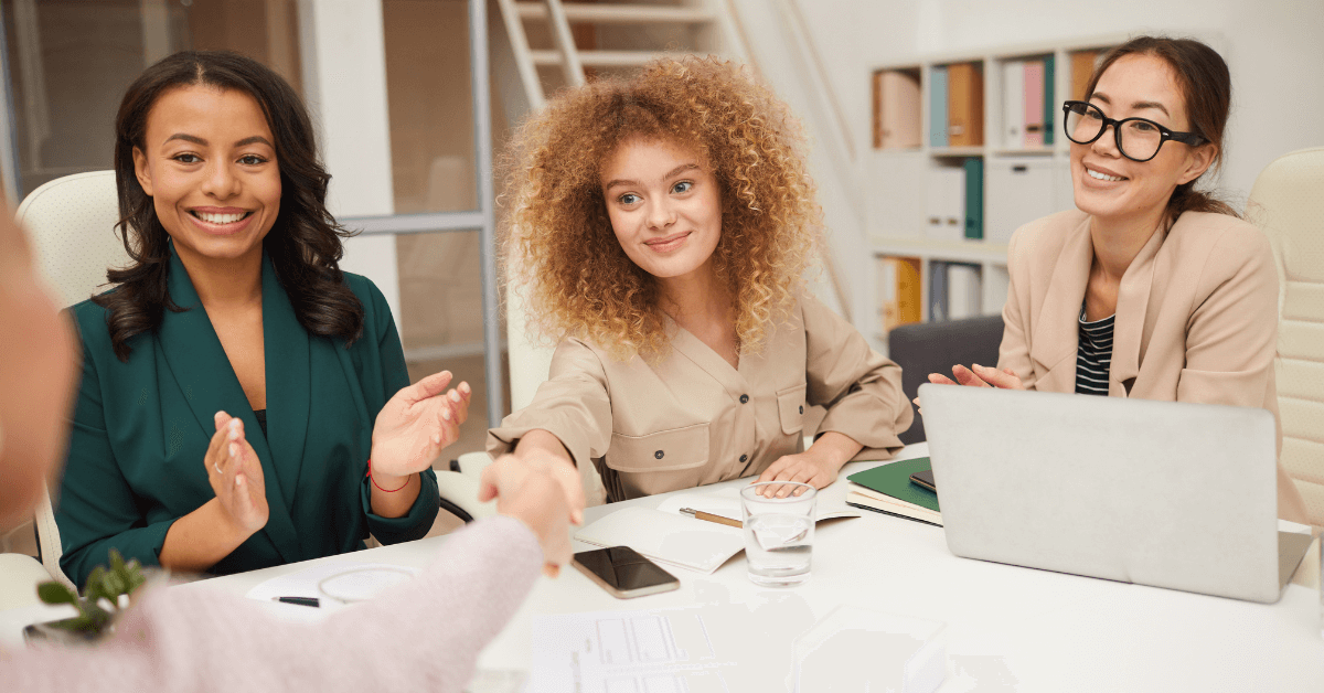 Three women smiling and clapping during a friendly negotiation, illustrating a successful interaction in the context of learning how to haggle while traveling abroad.