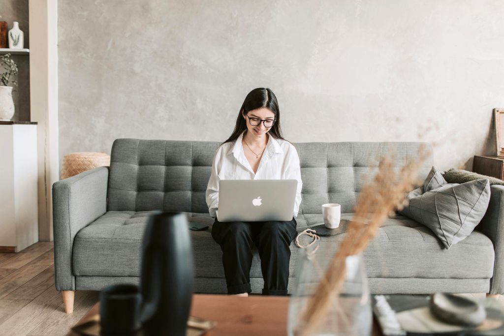 woman in glasses sits on a gray couch with a laptop, smiling while working. Researching how to haggle respectfully before traveling can prepare you for successful negotiations. Avoiding overpaying means more budget for experiences.