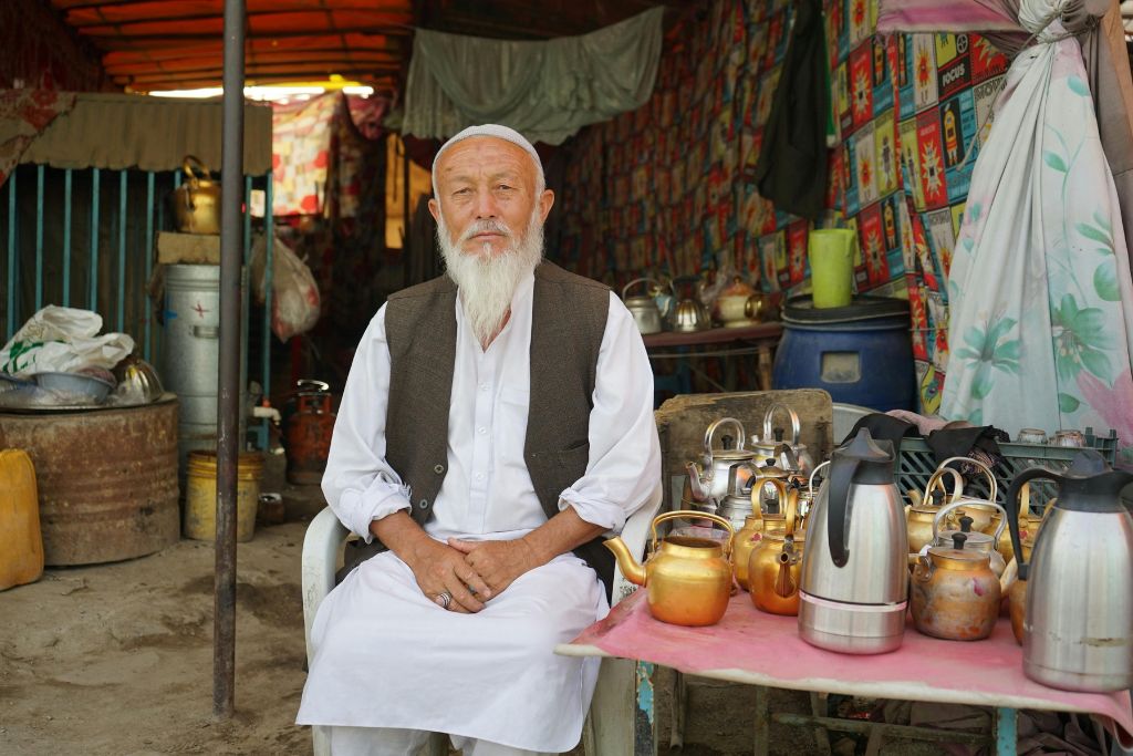 n elderly man with a white beard sits behind a table of copper and brass teapots in a market stall. Learning how to haggle while traveling abroad can help you negotiate better in local markets. Vendors appreciate bargaining etiquette when done respectfully.