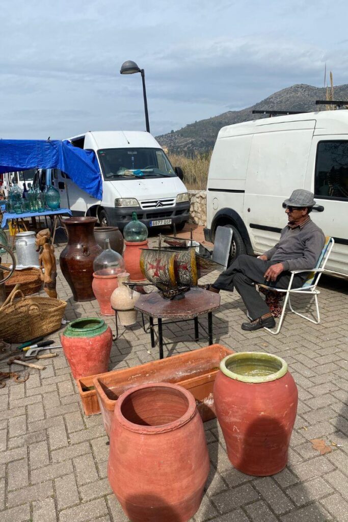 A vendor sits relaxed in a chair surrounded by pottery and antiques at an outdoor market. Learning how to haggle while traveling can help secure unique finds without overpaying. Always test your bargaining power before buying.