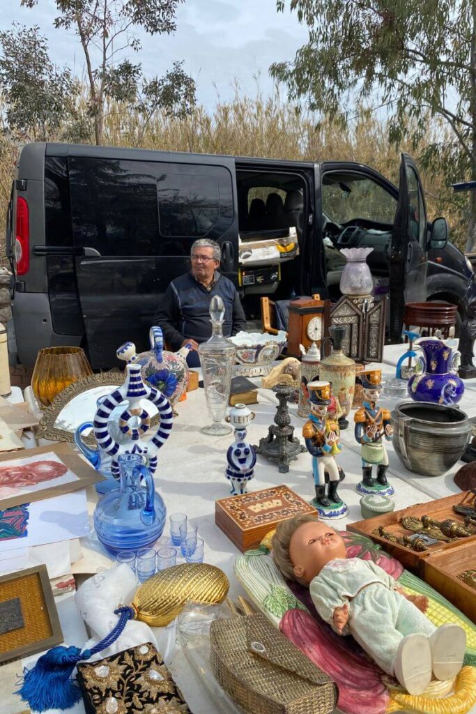 A vendor sits beside a table full of antiques, glassware, and collectibles, with an open van behind him. A perfect setting for haggling in Marrakech or any open-air market. Knowing how to bargain while on vacation can help you find hidden gems at a great price.