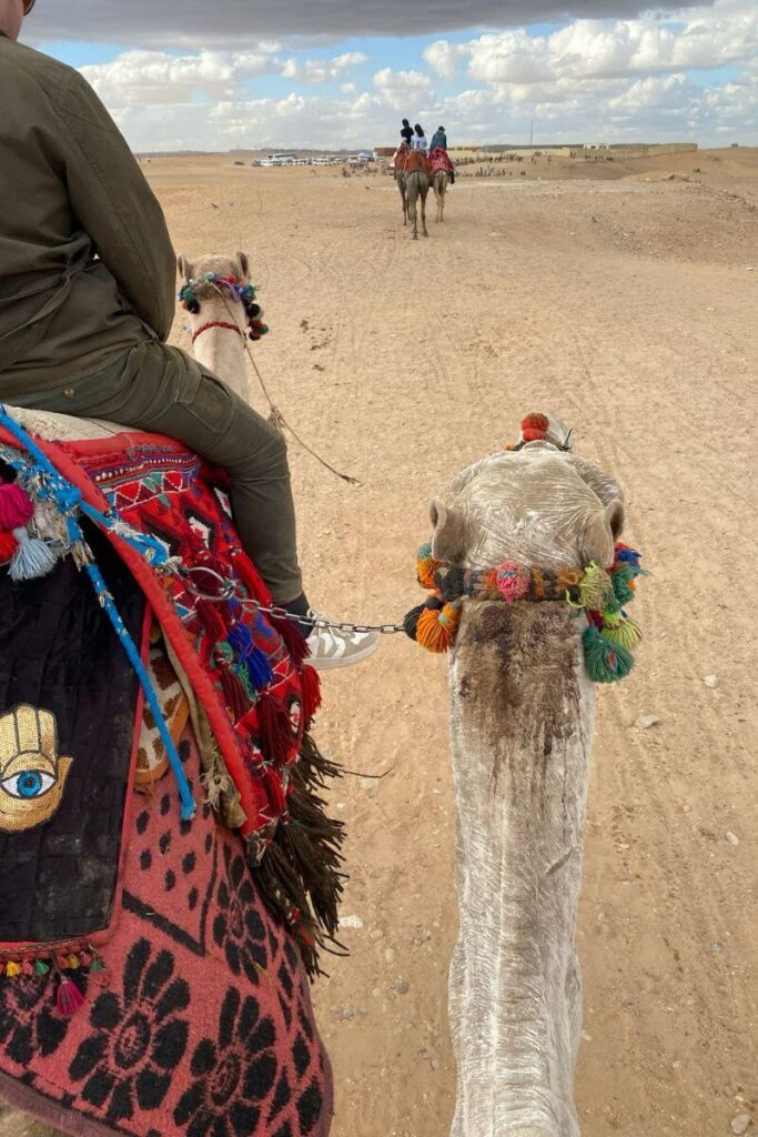 A first-person perspective of a camel ride through the desert near the pyramids. The camel’s back is adorned with a colorful saddle, and other travelers on camels are seen ahead. Many tourists opt for a tour guide for Giza Pyramids to enhance their experience while visiting pyramids of Egypt.