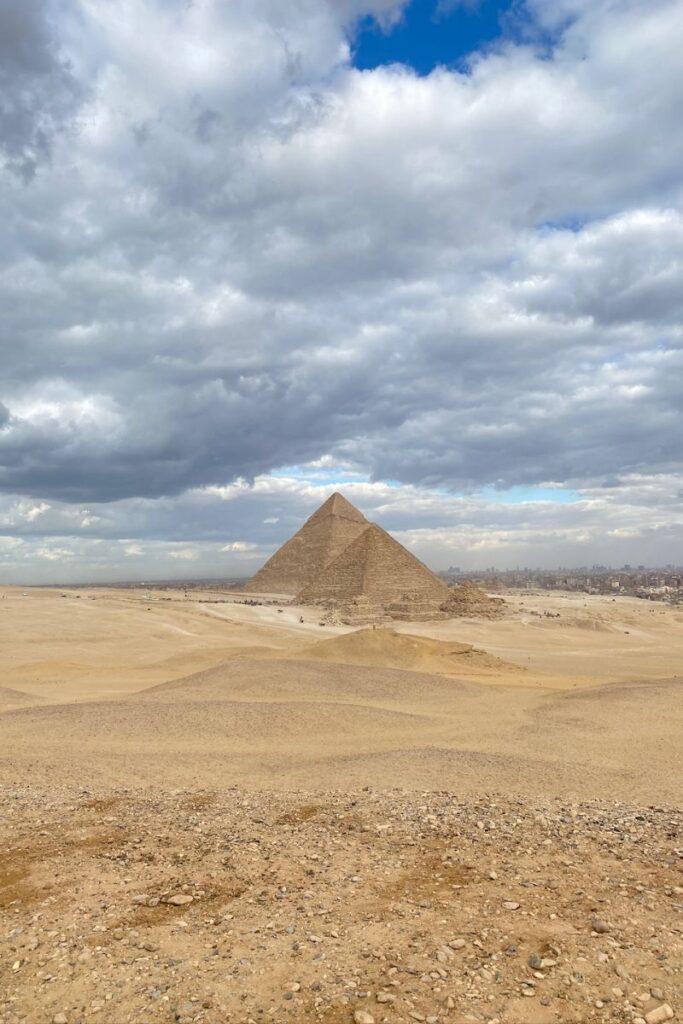 A distant view of the Great Pyramid of Giza under a cloudy sky, surrounded by desert. The pyramid stands tall with the vast sandy landscape stretching around it. Visiting the Great Pyramids of Giza is a must for travelers wanting to experience the Pyramids of Giza reality.