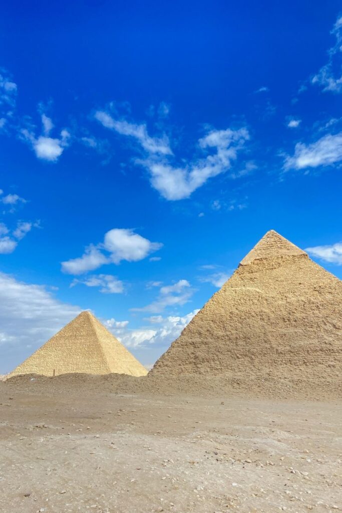 A striking shot of two pyramids with a bright blue sky and scattered white clouds above. The towering structures demonstrate the Pyramids of Giza height, a fascinating topic for those interested in who built the Pyramids of Giza.