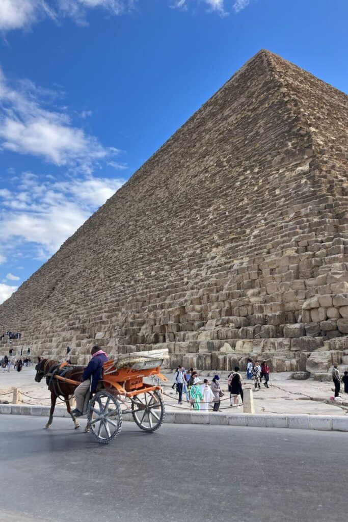 A horse-drawn carriage passes by the massive stone base of the Great Pyramid of Giza. Tourists walk along the stone path, exploring the site. Many visitors wonder, can you climb the Giza Pyramids, but access is restricted to preserve the site.