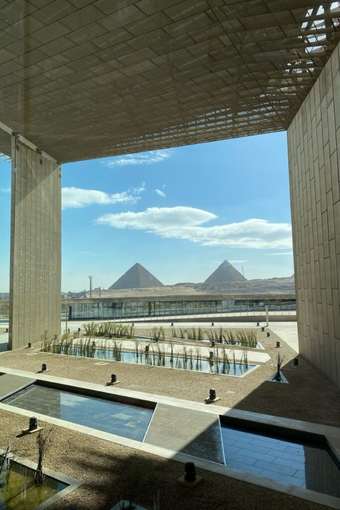 A breathtaking view of the Grand Egyptian Museum and pyramids, framed by modern architecture, with landscaped pools and greenery leading toward the Pyramids of Giza, showing the Grand Egyptian Museum location.