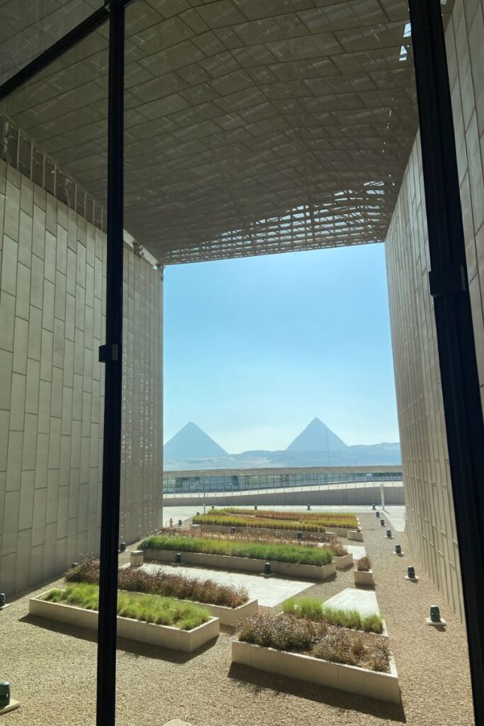 A scenic perspective of the Grand Egyptian Museum and pyramids, seen through a massive glass structure, highlighting the museum's strategic Grand Egyptian Museum location near the Great Pyramids of Giza.