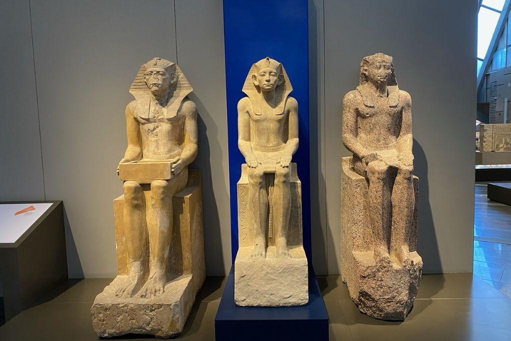 Three seated statues of ancient Egyptian figures displayed inside the Grand Egyptian Museum, highlighting the diversity of styles and materials—an unmissable part of things to see at the Grand Egyptian Museum.