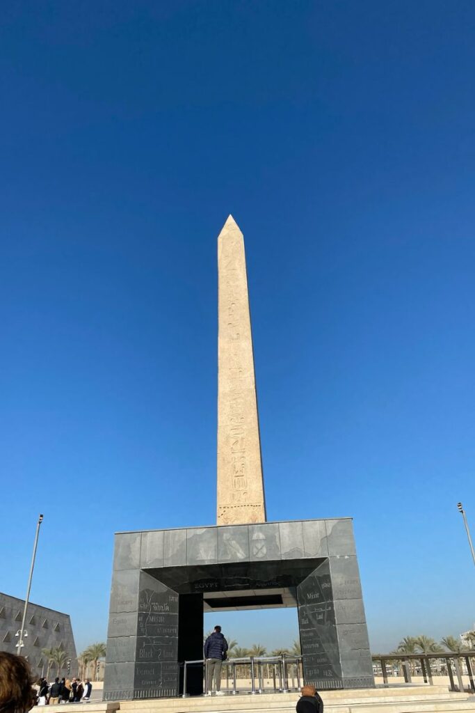 A towering obelisk with hieroglyphic inscriptions stands above the entrance of the Cairo Grand Egyptian Museum, showcasing the Grand Egyptian Museum architecture against a bright blue sky.