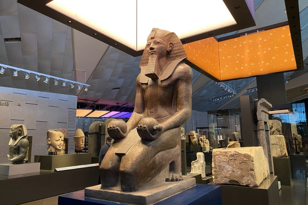 A seated statue of an Egyptian pharaoh displayed under a modern lighting installation in the Grand Egyptian Museum, highlighting the fusion of Grand Egyptian Museum architecture and ancient history.