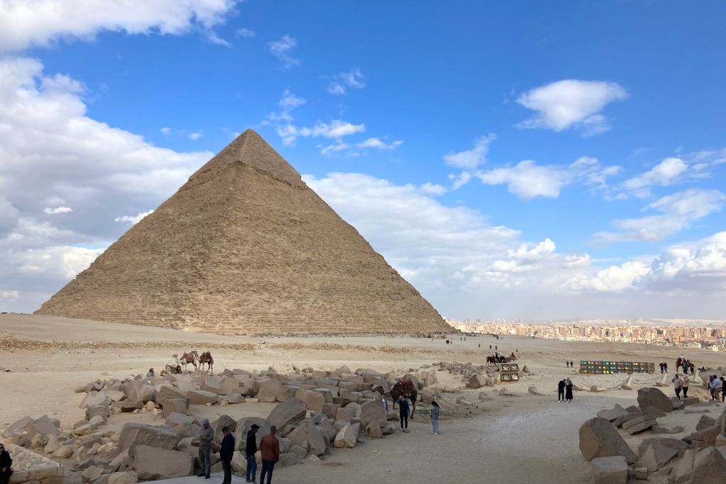 The Great Pyramid of Giza stands tall under a partly cloudy sky, with tourists exploring the site below. Understanding the Pyramids of Giza location helps plan the best itinerary when visiting the Great Pyramids of Giza.