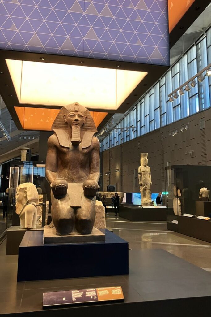 A massive seated statue of a pharaoh inside a modern museum, surrounded by other ancient artifacts. This display highlights Pyramids of Giza facts, offering insight into who built the Pyramids of Giza.