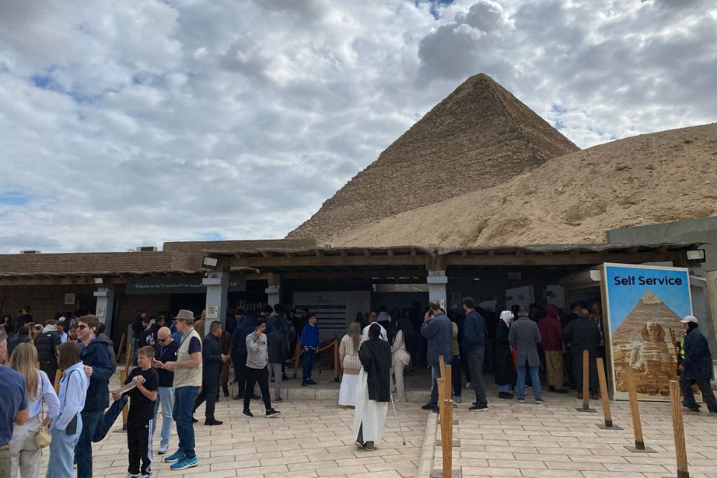 A crowded entrance area at the Pyramids of Giza, with tourists waiting in line under a partly cloudy sky. The Pyramids of Giza entrance fee is a key consideration for visitors, and checking the Giza Pyramids schedule in advance helps avoid long wait times.