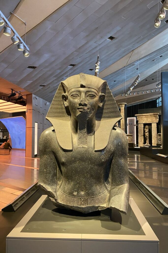 A massive bust of an ancient Egyptian pharaoh displayed inside the Grand Egyptian Museum Cairo, one of the biggest Egyptian museums, with an exhibition hall showcasing other artifacts in the background.