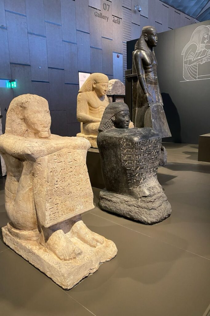 collection of ancient Egyptian seated statues, some holding inscribed stone slabs, displayed in the Grand Egyptian Museum Giza, a must-see attraction for those wondering what to see at the Grand Egyptian Museum.