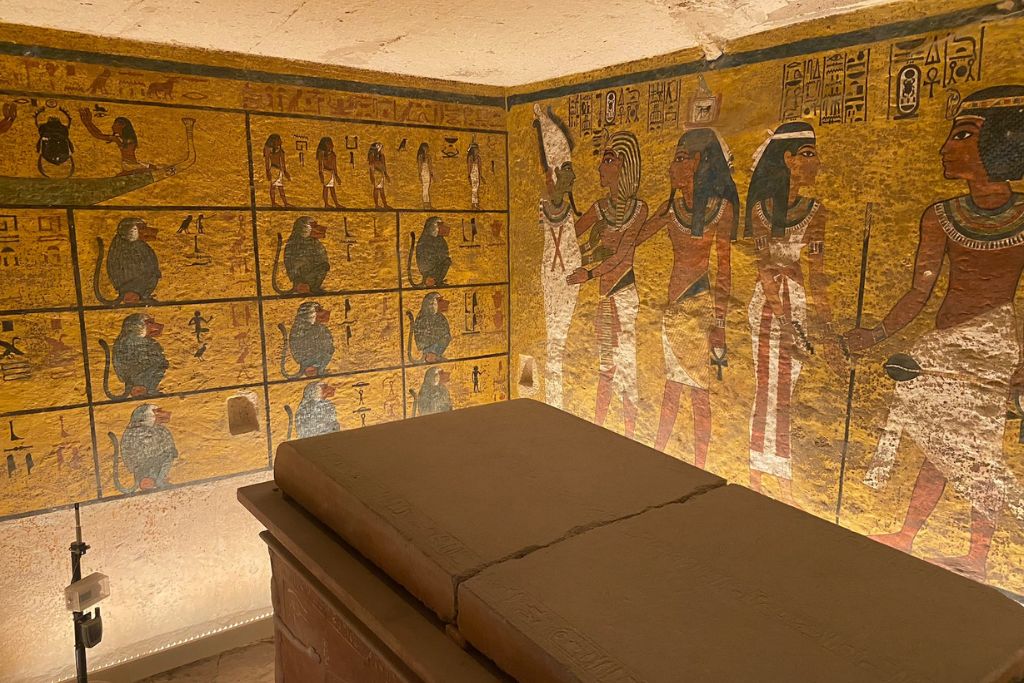 The interior of a royal Egyptian tomb, with colorful murals of ancient gods, baboons, and hieroglyphs surrounding a stone sarcophagus—one of the top Grand Egyptian Museum highlights for visitors.