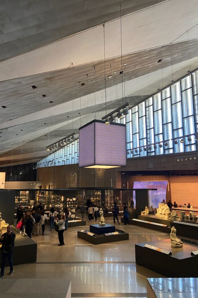 A spacious exhibition hall inside the Grand Egyptian Museum Cairo, filled with ancient artifacts and tourists, with modern hanging lights illuminating the displays—an essential stop for those planning a Grand Egyptian Museum guided tour.