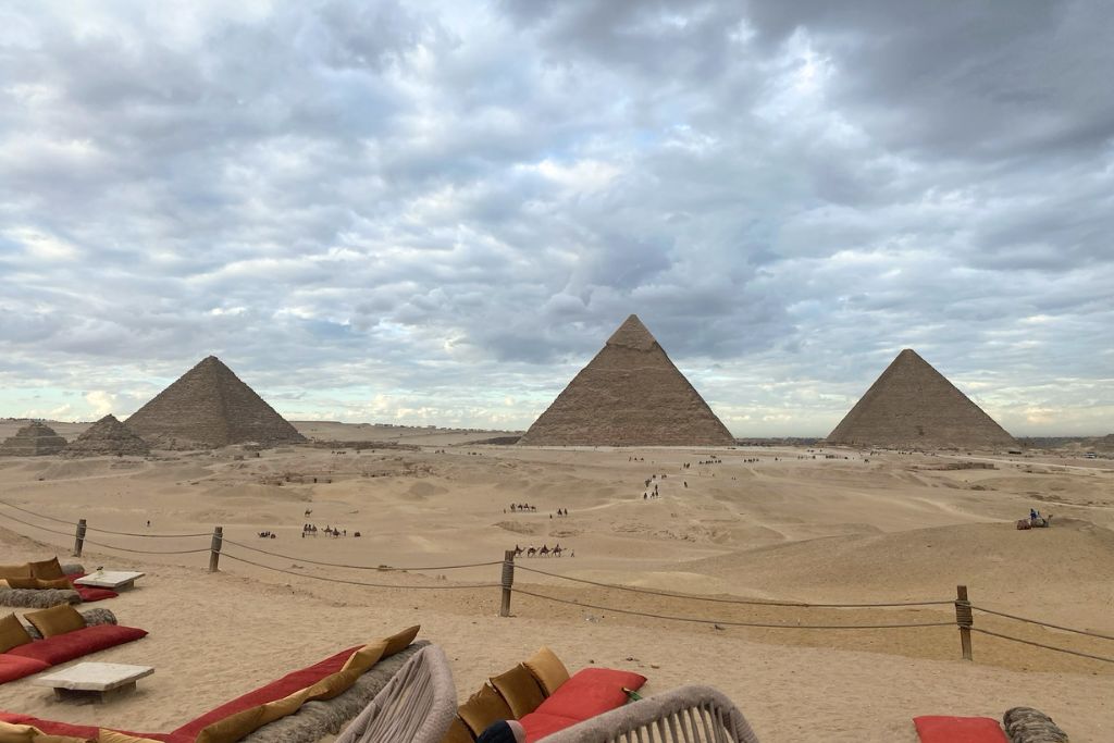 A cozy desert lounge setup with colorful cushions and a low wooden table, overlooking the three pyramids under a dramatic sky. This spot offers a relaxing Giza Pyramids view inn, making it an ideal location to enjoy the Pyramids of Giza worth visiting.