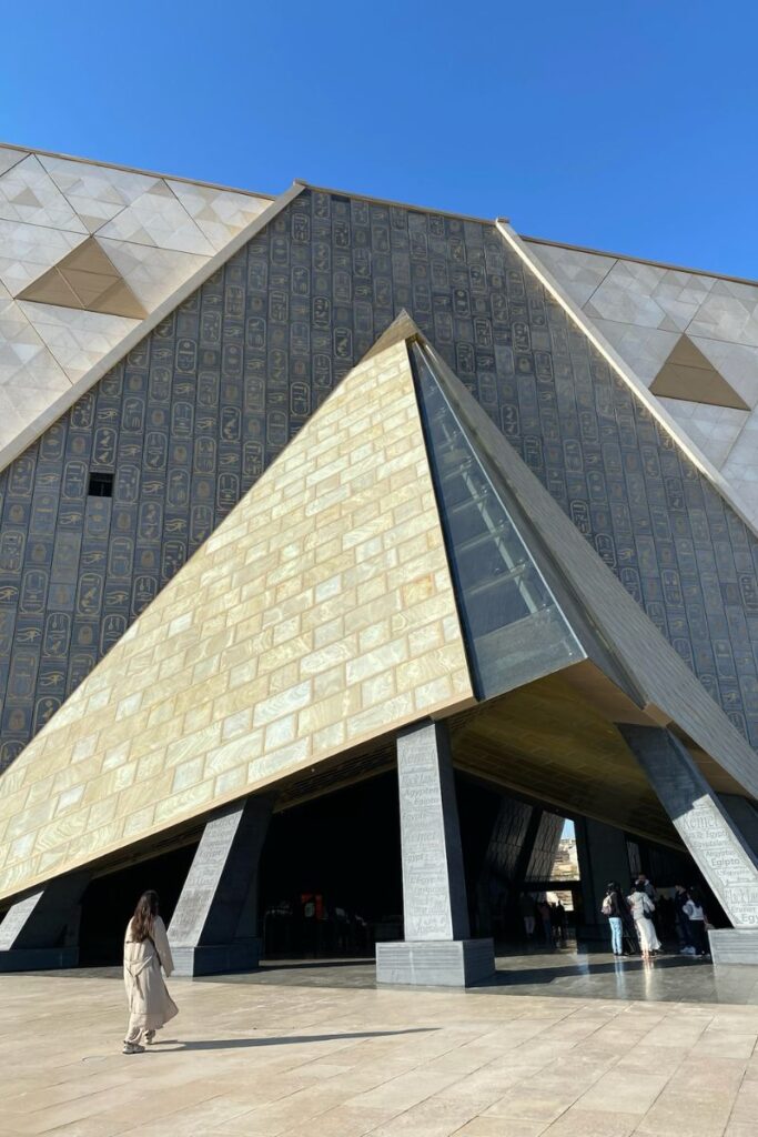 The Grand Egyptian Museum architecture, featuring a dramatic pyramid-inspired entrance adorned with hieroglyphic carvings, with visitors walking towards the Grand Egyptian Museum grand opening.