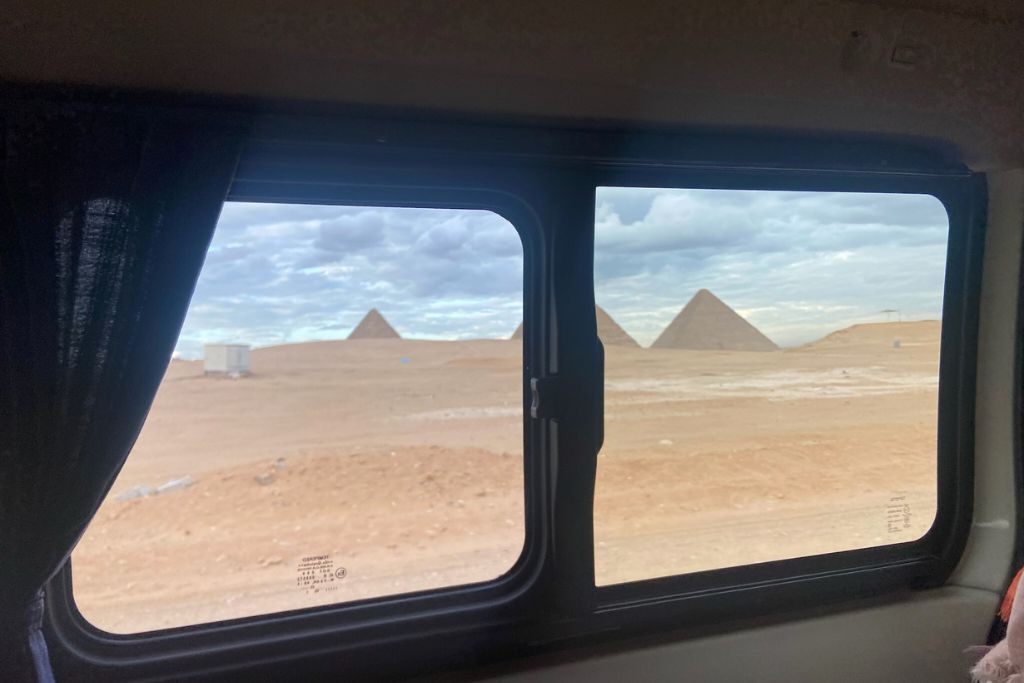 A framed view of the three pyramids from inside a vehicle, showing the desert landscape through the window. Arriving at the Pyramids of Giza Egypt early is one of the tips for visiting the Pyramids of Giza to avoid crowds.