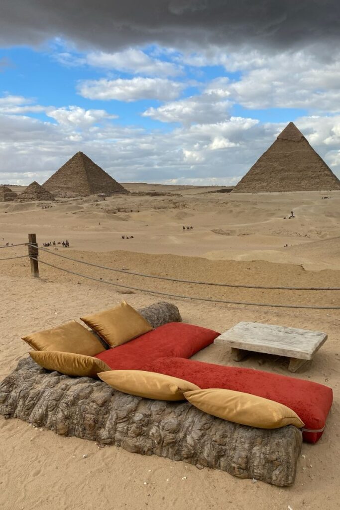 A panoramic view of the three pyramids of Giza under a cloudy sky, with seating in the foreground. The Giza Pyramids schedule allows visitors to explore these ancient structures, and the Pyramids of Giza official website provides updates on events and tours.