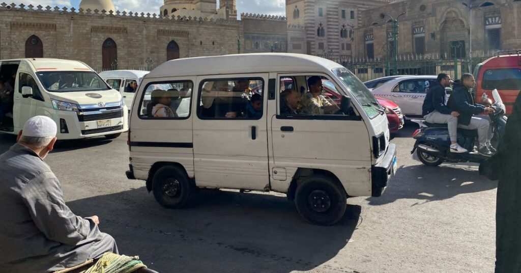 Busy Cairo street with a white minibus full of passengers, local Egyptian men, and motorbike riders in front of historic Islamic architecture. Shows traditional Egyptian taxi options versus using Uber Cairo or Uber in Egypt.