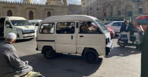 Busy Cairo street with a white minibus full of passengers, local Egyptian men, and motorbike riders in front of historic Islamic architecture. Shows traditional Egyptian taxi options versus using Uber Cairo or Uber in Egypt.