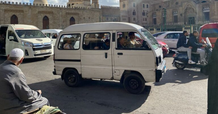 Busy Cairo street with a white minibus full of passengers, local Egyptian men, and motorbike riders in front of historic Islamic architecture. Shows traditional Egyptian taxi options versus using Uber Cairo or Uber in Egypt.