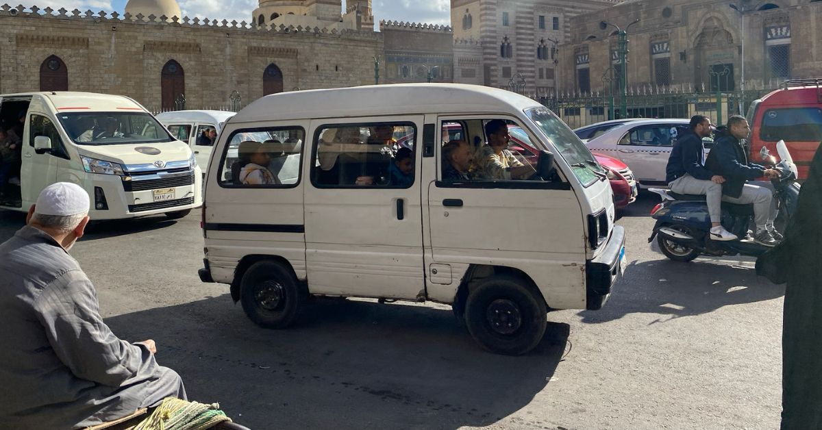 Busy Cairo street with a white minibus full of passengers, local Egyptian men, and motorbike riders in front of historic Islamic architecture. Shows traditional Egyptian taxi options versus using Uber Cairo or Uber in Egypt.