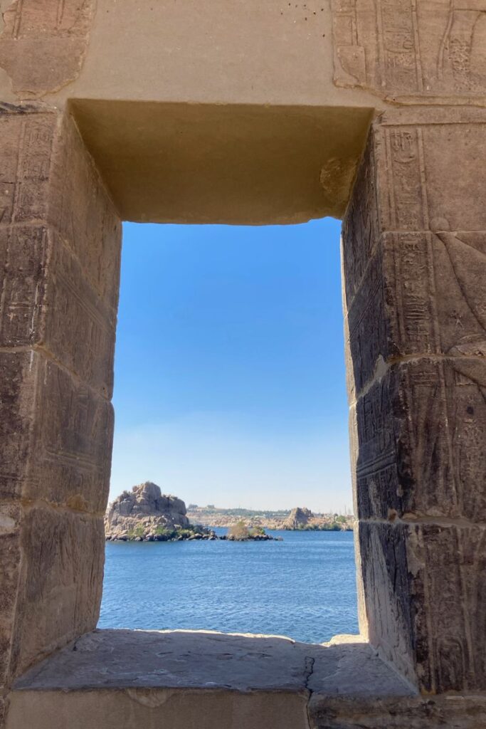 View through a stone window at the Temple of Isis on Philae Island, Egypt, showing the sparkling Nile River and rock formations in the distance, a peaceful glimpse from within the ancient temple.
