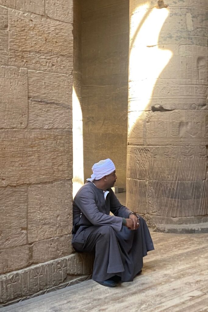 A local man in traditional dress rests beside a hieroglyph-covered column in the Temple of Isis on Philae Island, Egypt, where ancient stonework and natural light create a serene atmosphere.